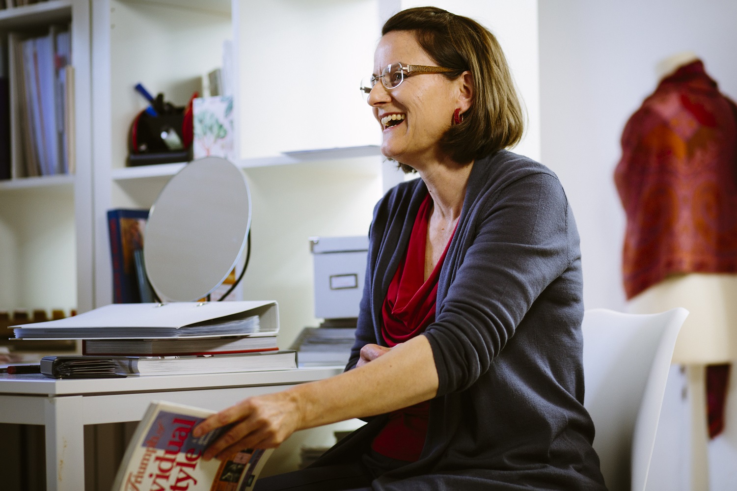 Mittelalte Frau sitzt lachend auf einem Stuhl. Auf ihrem Schoß liegt ein aufgeschlagenes Buch. Im Hintergrund sieht man ein Regal, auf dem weitere Bücher und Ordner stehen.
