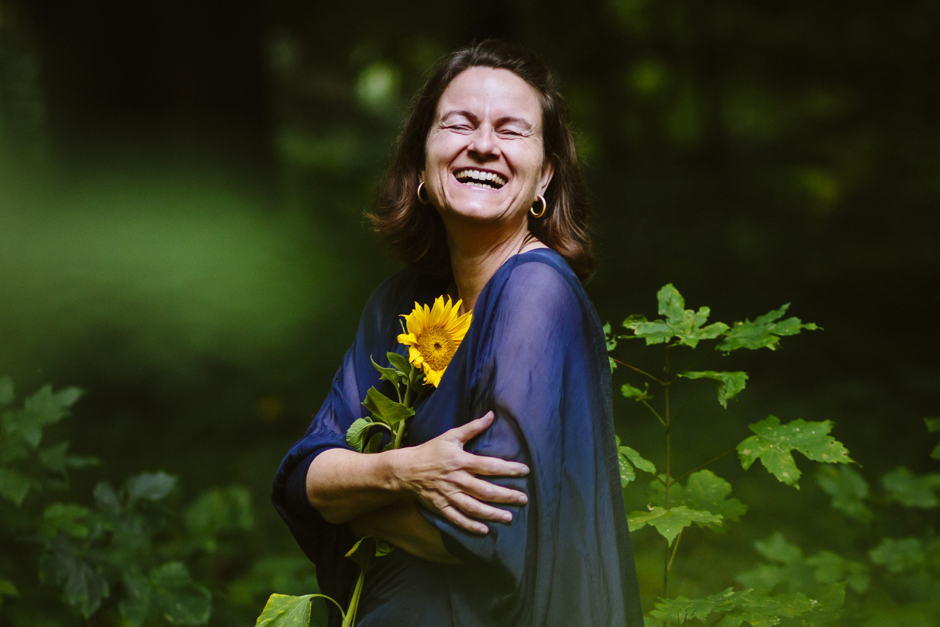 Mittelalte Frau, die im Wald steht und in die Kamera blickt, sie lacht mit weit geöffnetem Mund ganz entspannt. Sie trägt ein blaues Oberteil und hat eine Sonnenblume im Arm.