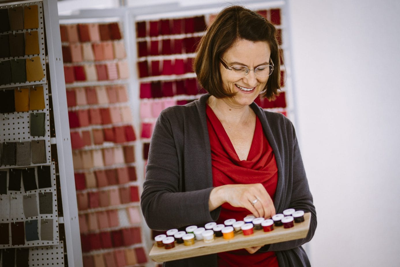 Frau in rotem Oberteil hält ein Holztablett in der Hand und schaut darauf. Auf dem Tablett sind kleine Töpfchen angeordnet, die verschiedene Acrylfarben enthalten.