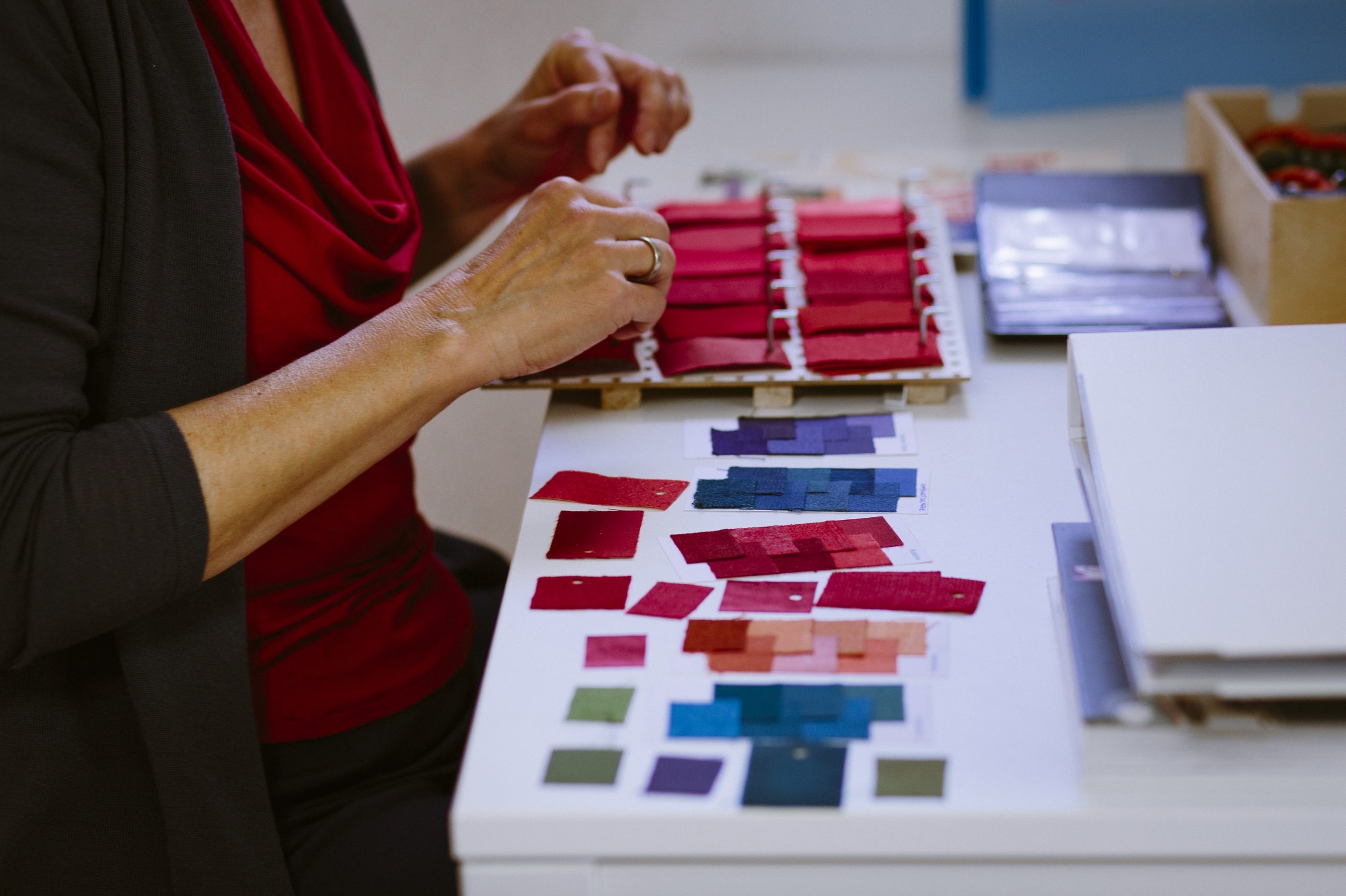 Eine Frau sitzt vor einem Tisch, auf dem vschiedenen kleine Stoffstücke liegen. Man sieht das Gesicht der Frau nicht, nur ihre Unterarme und ihre Hände. Die Hände scheinen über dem Tisch zu schweben. Im hinteren Teil des Tischs liegt ein Holzbrett, auf dem vschiedene rote kleine Stoffstücke aufgepinnt sind.