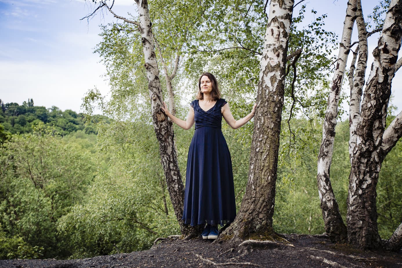 Es sind vier schmale Bäume auf dem Bild zu sehen, dahinter die Wipfel von weiteren Bäumen. Eine mittelalte Frau in einem dunkelblauen langen Kleid steht zwischen zwei Bäumen und hat die Hände auf Ellenbogenhöhe an den beiden Baumstämmen.
