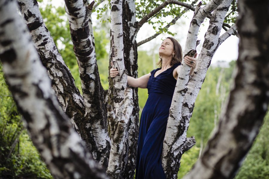 Stefanie steht mit zurückgeneigtem Kopf und ernstem Gesichtsausdruck da und hält sich an zwei schmalen Bäumen fest.