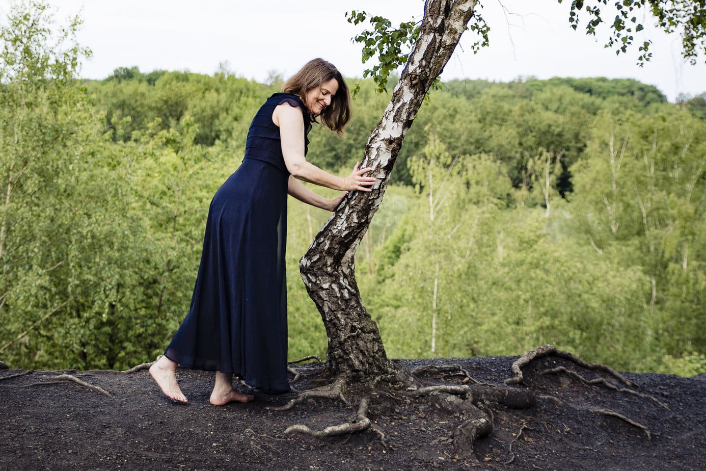 Mittelalte Frau in langem blauen Kleid steht neben einem Baum. Sie lächelt. Es sieht so aus, als würde sie mit dem Baum sprechen oder tanzen.
