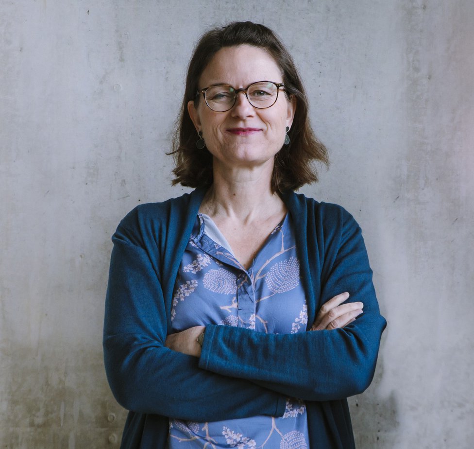 Eine mittelalte Frau mit halblangen dunkelbraunen Haaren und Brille steht vor einer hellen Betonwand. Sie lächelt mit geschlossenem Mund und hat die Arme verschränkt.