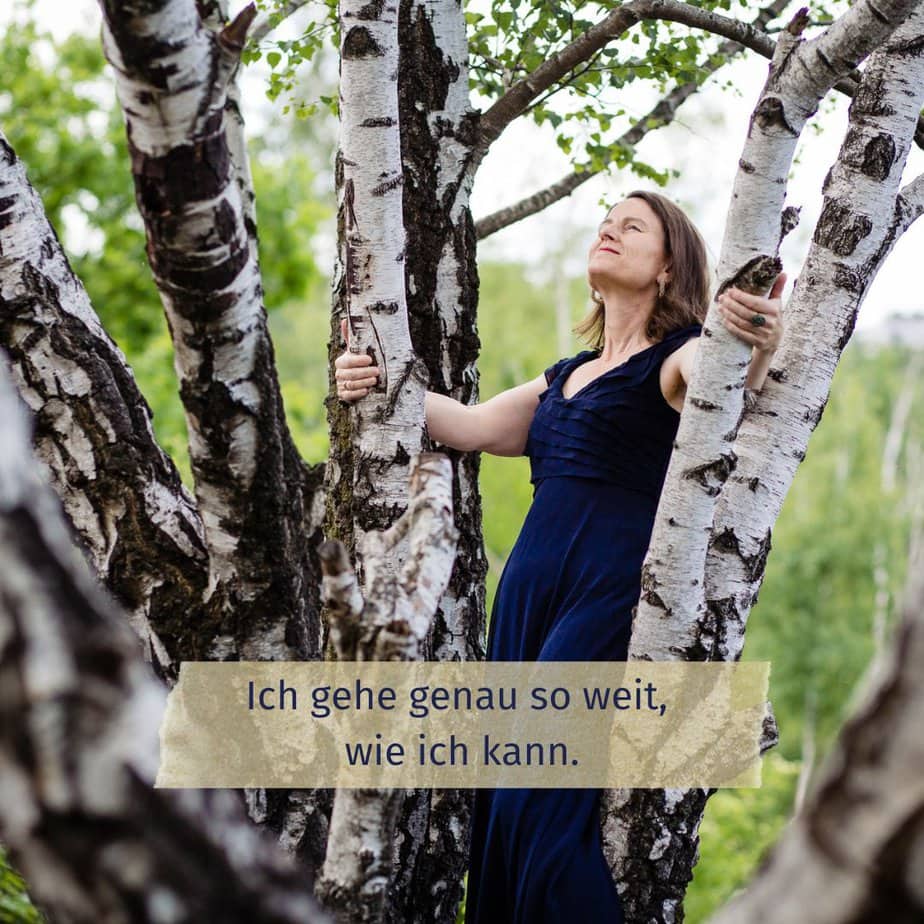 Stefanie Sauer-Bloch steht hinter einigen Birken und hält sich an zwei von ihnen fest. Sie lehnt sich etwas nach hinten und blickt in den Himmel.
Unten auf dem Foto steh der Satz: "Ich gehe genau so weit, wie ich kann."